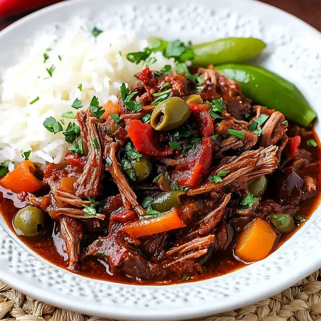 A plate of shredded beef stew garnished with herbs, olives, and bell peppers, served alongside white rice and green snap peas.