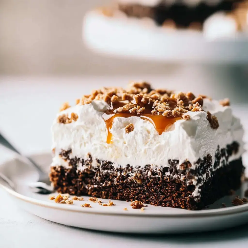 A slice of chocolate cake layered with whipped cream and drizzled with caramel, topped with crushed cookies, on a white plate.