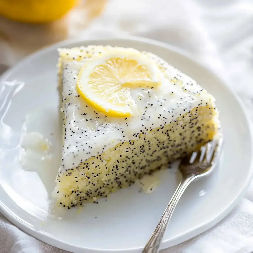 A slice of lemon poppy seed cake topped with a creamy frosting and a slice of lemon is displayed on a white plate.