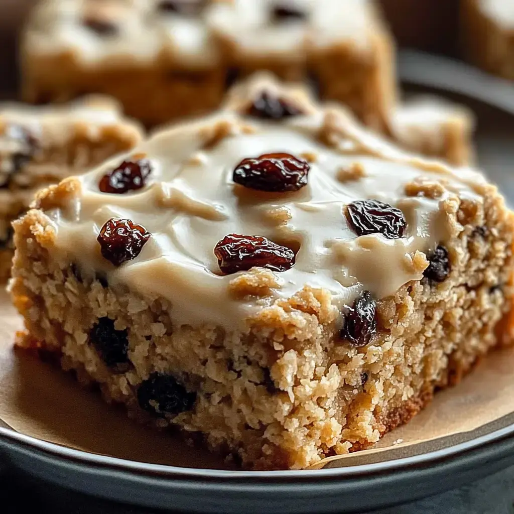 A close-up of a slice of cake topped with a creamy icing and dotted with raisins, resting on a plate.