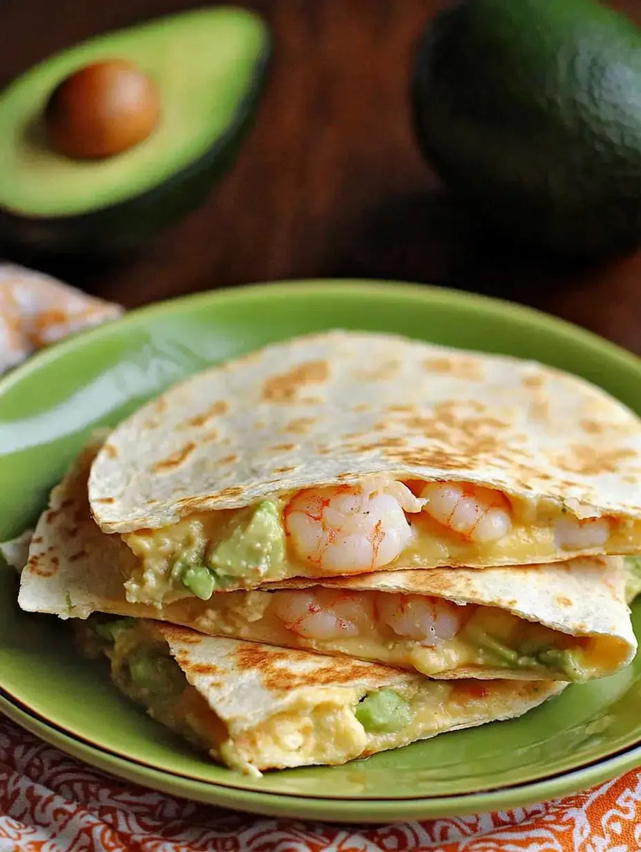 A close-up of a green plate featuring stacked quesadillas filled with shrimp and avocado, accompanied by a halved avocado in the background.