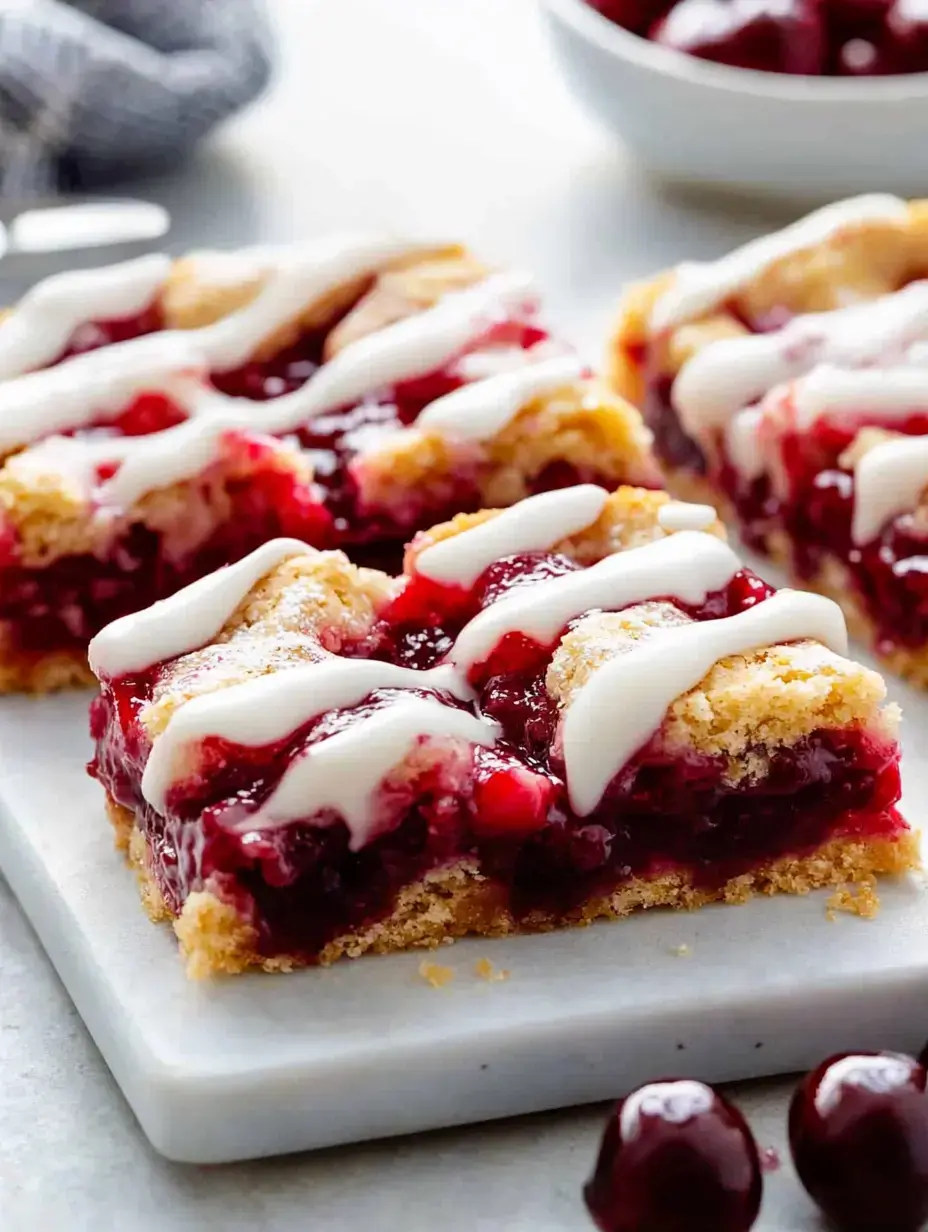 A plate of cherry-studded dessert bars drizzled with white icing, with a few whole cherries nearby.