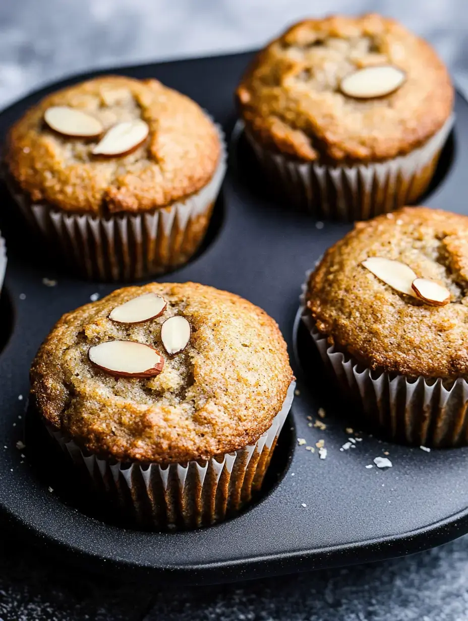 Four freshly baked almond muffins are placed in a black muffin tray with slivered almonds on top.