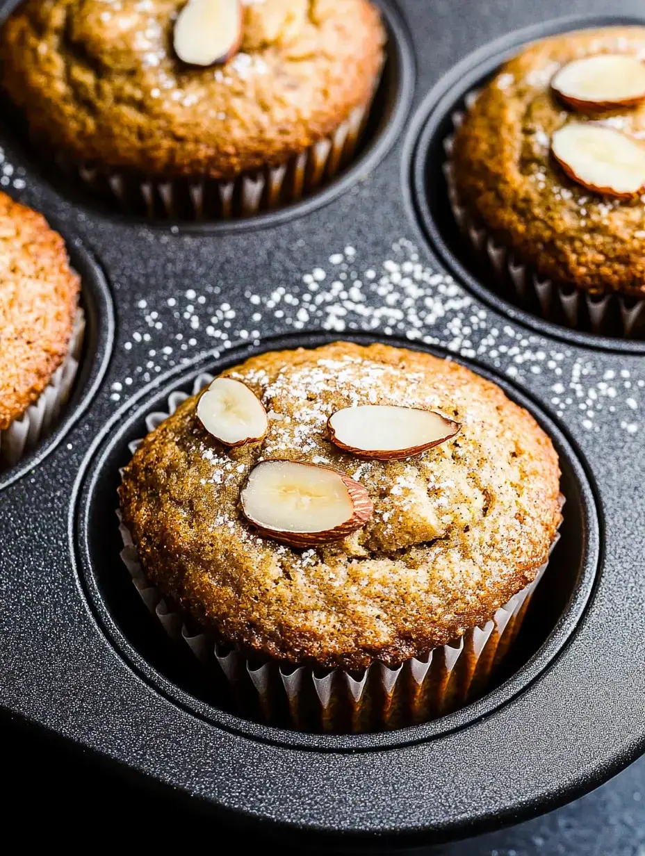 A close-up of freshly baked muffins topped with sliced almonds and powdered sugar, set in a dark muffin tin.