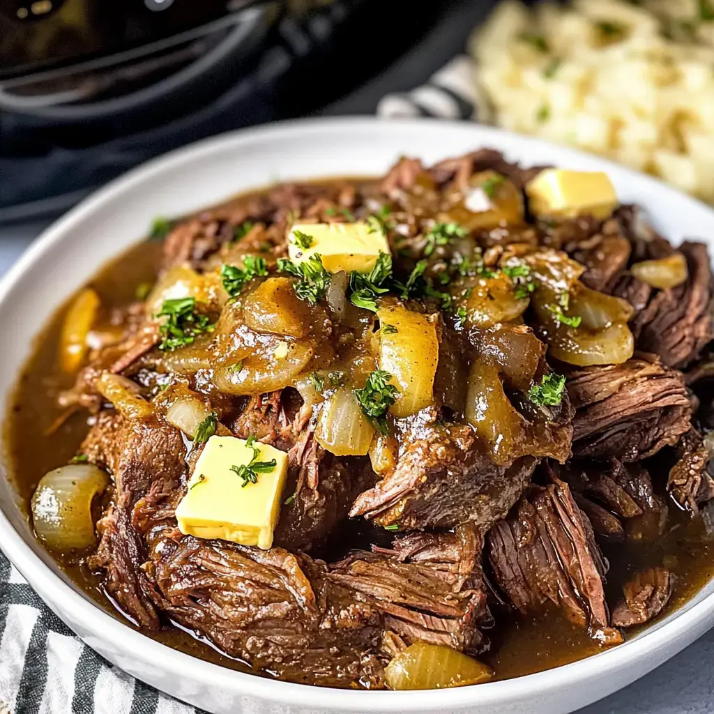 A close-up of a bowl of tender beef roast topped with onions, gravy, butter cubes, and parsley, accompanied by a side of mashed potatoes.