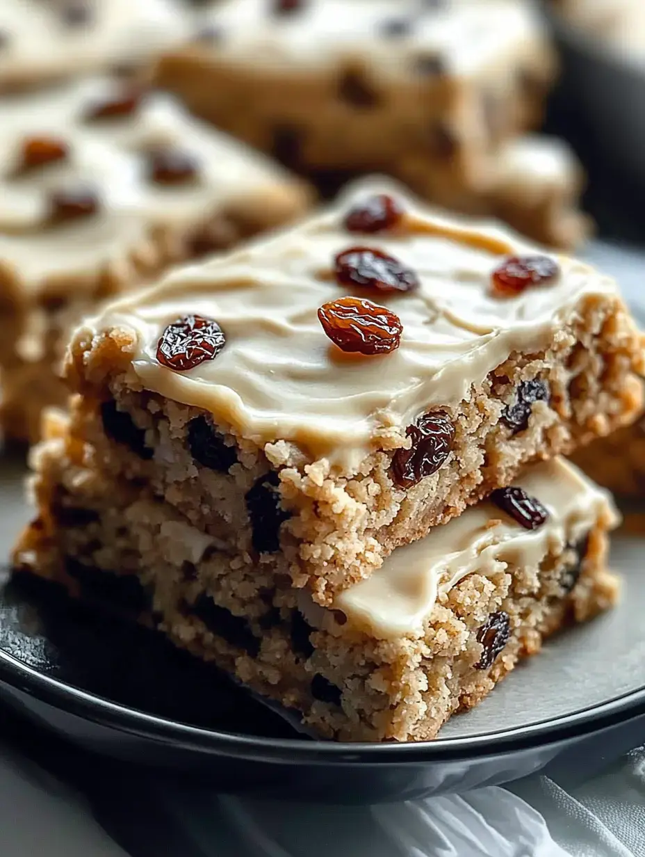 A close-up of two stacked pieces of frosted cake with raisins on top, displayed on a black plate.