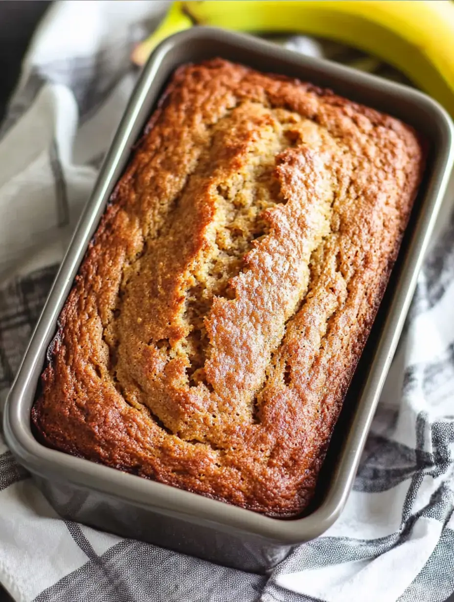 A freshly baked loaf of banana bread sits in a gray baking pan, accompanied by ripe bananas and a patterned cloth.