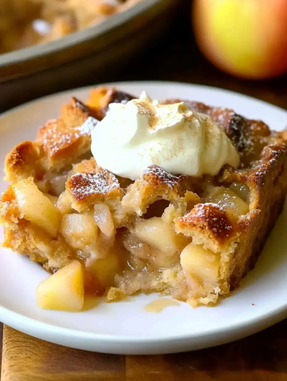 A slice of apple pie is served on a plate, topped with whipped cream and dusted with powdered sugar, with a whole apple in the background.