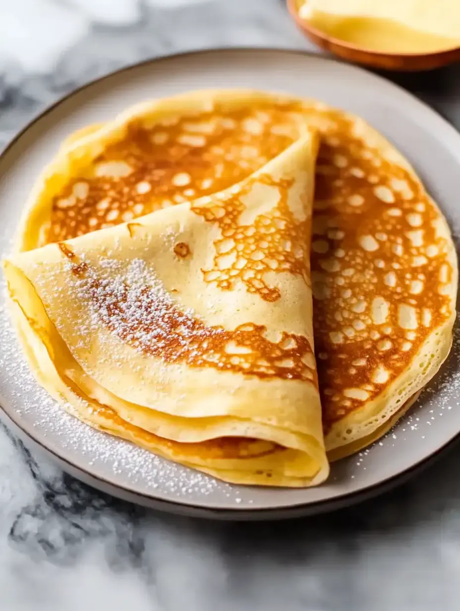 A plate of freshly made golden-brown crepes dusted with powdered sugar.