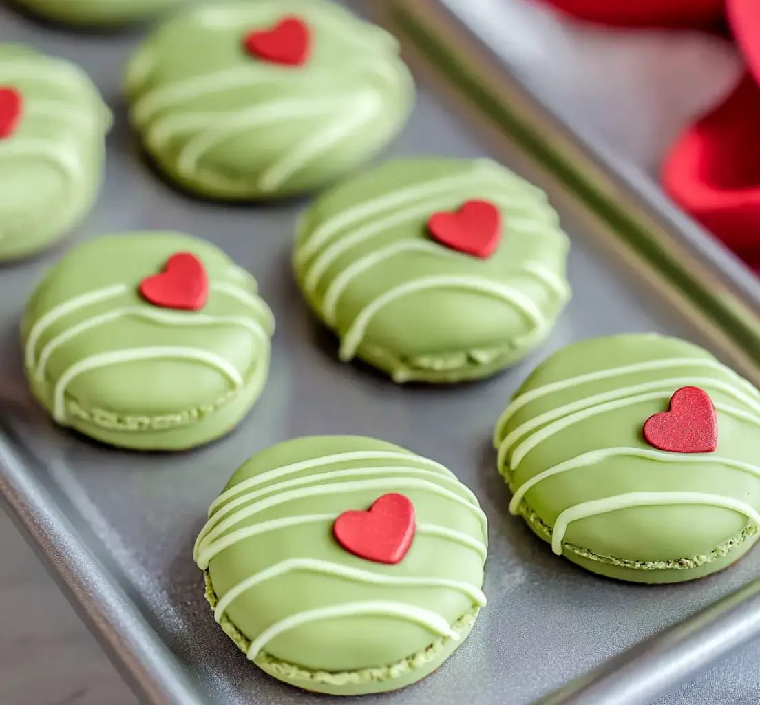 A tray of green macarons decorated with red heart toppings and white drizzle.