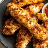 A close-up image of golden-brown breaded chicken strips served on a plate with a side of dipping sauce.