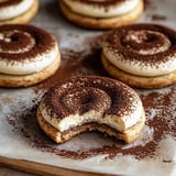 A close-up of a cookie with a bite taken out, showing a creamy filling and topped with a dusting of cocoa powder, surrounded by more similar cookies on a baking sheet.