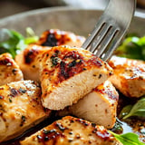 A close-up of tender, grilled chicken pieces garnished with herbs, with a fork piercing one of the pieces.