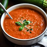 A bowl of red soup garnished with fresh basil leaves sits on a wooden surface, accompanied by zucchini in the background.