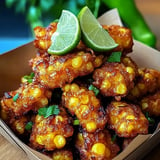 A close-up of crispy corn fritters garnished with lime wedges and fresh herbs in a brown container.