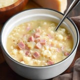 A close-up of a bowl of hearty soup containing diced ham, cauliflower, and vegetables, with a spoon resting inside.