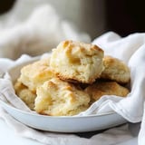 A pile of golden-brown biscuits arranged in a bowl, with one half-biscuit prominently placed in front, resting on a white cloth.