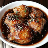 A white dish filled with tender, glazed chicken thighs covered in dark sauce, garnished with sesame seeds and chopped herbs.