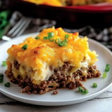 A slice of cheesy shepherd's pie with ground beef and mashed potatoes, garnished with chopped green onions, on a white plate.