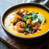 A bowl of creamy orange soup topped with shrimp, crispy bits, cilantro, sliced jalapeños, and a lime wedge.