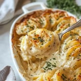 A spoon is lifting a creamy, cheesy, and baked potato dish from a white casserole, garnished with herbs.