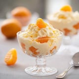 A glass dessert dish filled with whipped cream and orange segments, topped with a slice of orange, is displayed alongside whole oranges.