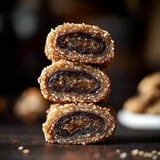 Three stacked pieces of sweet, rolled dates covered in a coarse, light-colored sugar are displayed against a blurred background.