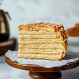 A slice of layered pastry dessert with cream filling is displayed on a wooden cake stand.